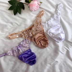 Può includere: Tre paia di biancheria intima in beige, bianco e viola, ciascuna caratterizzata da una base in raso e un pannello in pizzo trasparente con ricami floreali. I capi includono spalline regolabili e piccoli fiocchi al centro della vita.