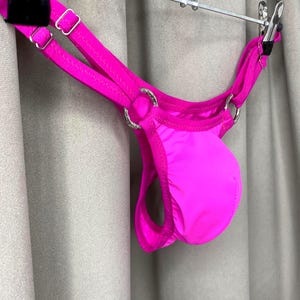 May include: Bright pink thong-style underwear with silver-colored metal rings and straps. The garment is suspended from a metal hanger with a black clip. The fabric has a smooth, shiny appearance.