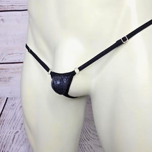 May include: A black thong featuring a small, dark blue textured pouch with a subtle speckled pattern. The garment is constructed with thin, adjustable black straps connected by small silver-toned metal rings on each side of the front pouch.