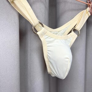Op de afbeelding: Ivoor kleurige string met een pouch design. Het kledingstuk heeft een zachte, gladde stof en wordt bij elkaar gehouden door crèmekleurige bandjes en zilveren metalen ringen. Het ondergoed hangt voor een grijze gordijn.