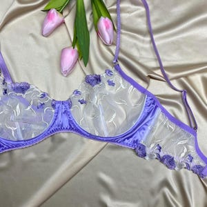 Può includere: Un reggiseno con ferretto color lavanda con coppe in rete bianca trasparente, ricami floreali viola, bordi in raso e spalline regolabili, presentato accanto a tre tulipani rosa chiaro su un tessuto di raso color champagne.
