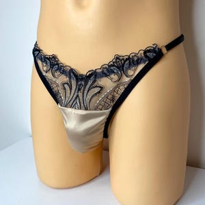 Könnte beinhalten: Ein beiger Satin-Tanga mit schwarzen und marineblauen Spitzendetails. Der Spitzensaum weist ein kompliziertes Blumenmuster auf. Der Tanga hat einen schwarzen Samtbund und eine bequeme Passform.