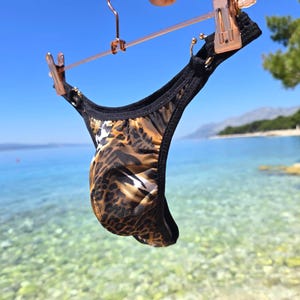 Op de afbeelding: Een onderbroek met een bruin en zwart luipaardprintpatroon met effen zwarte randen en afwerkingen. Het kledingstuk hangt aan een roségouden metalen hanger met twee clips, waardoor de textuur van de stof en het geplooide ontwerp zichtbaar zijn.