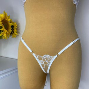 Op de afbeelding: Witte lingerie set met een bloemen kanten design. De string-stijl onderkant heeft delicate bandjes en een bloemen kanten detail. Deels zichtbare top heeft een vergelijkbaar bloemen kanten design. Een gele zonnebloem staat op de achtergrond.