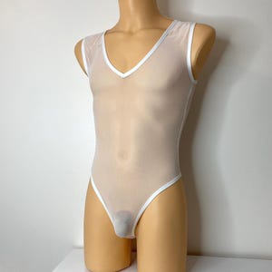 Puede incluir: Un body transparente sin mangas en color beige claro con ribete blanco en los bordes y escote en V. La prenda está diseñada para ser ajustada y tiene una pernera de corte alto.