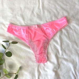 Könnte beinhalten: Hellrosa Satin-String-Unterwäsche mit einer kleinen Schleife vorne. Die Unterwäsche hat einen transparenten Einsatz in der Mitte und weiße Nähte an den Rändern. Der Stoff hat ein glänzendes Aussehen.