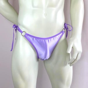 Pode incluir: Calcinha de biquíni lilás acetinada com laços e argolas de metal prateado. As bordas são aparadas com um tom roxo mais escuro. Um design simples e elegante para trajes de banho.