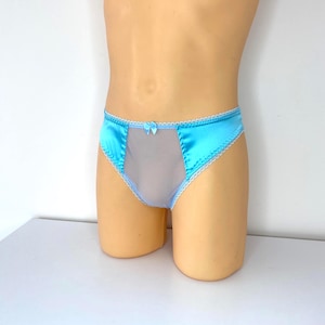 Puede incluir: Ropa interior de satén azul claro con un panel transparente en el centro. La ropa interior tiene un delicado ribete de encaje blanco a lo largo de los bordes y un pequeño lazo en la parte delantera. La prenda se exhibe en un maniquí.