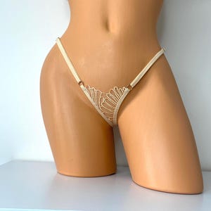 May include: Ivory-colored thong underwear with a delicate lace design on the front. The thong features thin straps and small metal rings at the sides. The lace has a fan-like pattern.