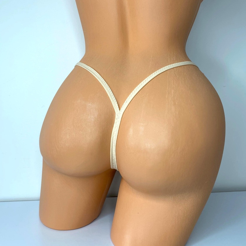 Op de afbeelding: Cr&egrave;mekleurig string-ondergoed. De string heeft dunne bandjes die een Y-vorm vormen aan de voor- en achterkant. Het ondergoed wordt getoond op een mannequin, die het ontwerp en de pasvorm van het kledingstuk laat zien. De achtergrond is lichtblauw.