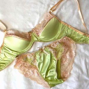 May include: A light green satin lingerie set, comprising a bra and matching briefs. Both pieces are trimmed with peach-coloured lace and small bows. The bra features adjustable straps. The set is presented on a white surface.