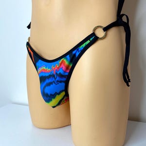 Puede incluir: Un tanga de traje de baño estampado con un diseño vibrante en azul, rojo y amarillo. Los bordes están ribeteados en negro y presenta anillos dorados y cierres de lazo negros. El traje de baño se exhibe en un maniquí.