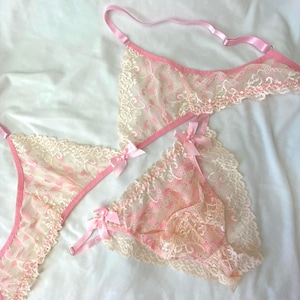 Könnte beinhalten: Ein dreiteiliges Dessous-Set in zartem Rosa und Creme. Das Set beinhaltet ein Bralette, einen String-Tanga und einen hoch taillierten Slip, alle mit zarter Spitze und kleinen rosa Schleifen verziert. Die Dessous werden auf einer weißen Oberfläche präsentiert.