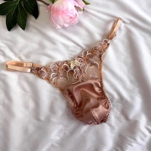 Può includere: Un perizoma con pannello frontale in raso color oro rosa e pannelli laterali in rete trasparente con intricati ricami floreali. Il capo presenta sottili spalline regolabili in raso e un piccolo fiocco decorativo al centro.