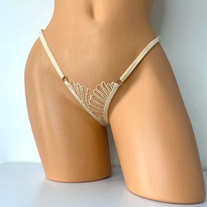 Op de afbeelding: Ivoor kleurige string met een delicaat kanten ontwerp aan de voorkant. De string heeft dunne bandjes en kleine metalen ringen. Het kant heeft een gedetailleerd, open patroon. Het kledingstuk wordt op een mannequin getoond.