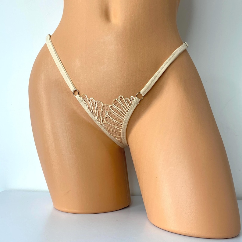 Op de afbeelding: Ivoor kleurige string met een delicaat kanten ontwerp aan de voorkant. De string heeft dunne bandjes en kleine metalen ringen. Het kant heeft een gedetailleerd, open patroon. Het kledingstuk wordt op een mannequin getoond.