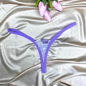 May include: A lavender thong featuring thin side straps and a central triangular panel made of sheer white lace with a delicate purple floral embroidery pattern. The garment is displayed against a smooth, light champagne-colored satin fabric.