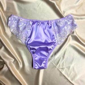 May include: Lilac satin underwear with delicate white floral lace trim. The lingerie features a high-cut design and small bow accents. The smooth, shiny fabric and intricate lace details create a feminine aesthetic.
