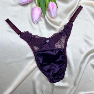 Può includere: Un perizoma viola scuro con un pannello frontale in raso e dettagli in pizzo intricati lungo la fascia in vita. Il capo presenta sottili spalline regolabili e un piccolo ciondolo argentato a forma di cuore al centro della parte in pizzo.