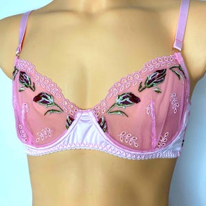 May include: A light pink underwire bra featuring sheer mesh cups with embroidered dark purple rosebud motifs and green stems. The garment includes scalloped edges, adjustable satin shoulder straps, and a solid pink satin base panel.