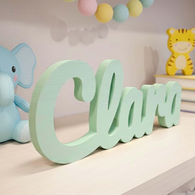 May include: A mint green wooden sign spells out the name "Clara" in a cursive font. The sign is placed on a light-colored shelf, with a blue elephant toy and a yellow tiger figurine in the background. A garland of pastel-colored felt balls hangs above.