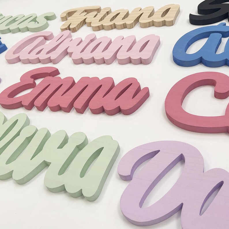 May include: A collection of wooden name cutouts in various colors, including pink, green, blue, and natural wood. The names are in a cursive font, with the name "Emma" prominently displayed in a vibrant pink. These decorative pieces are ideal for personalized room decor.