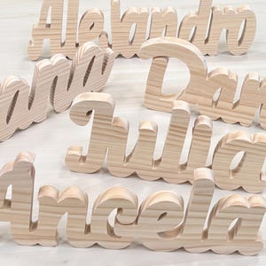 May include: Wooden name cutouts in a variety of names, including "Alejandro", "Julia", and "Angela". The names are in a cursive font and made of light-colored wood. Ideal for personalized decor or gifts.