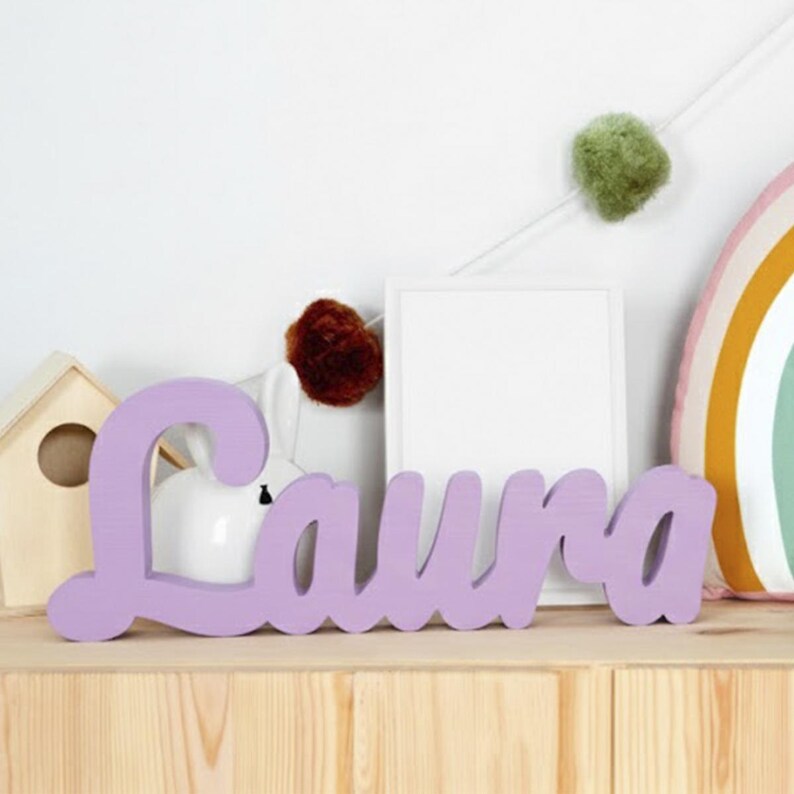 May include: A lavender-colored wooden sign spells out the name "Laura" in a cursive font. The sign is placed on a wooden shelf, with decorative items in the background, including a small birdhouse, a white frame, and a rainbow-shaped pillow.