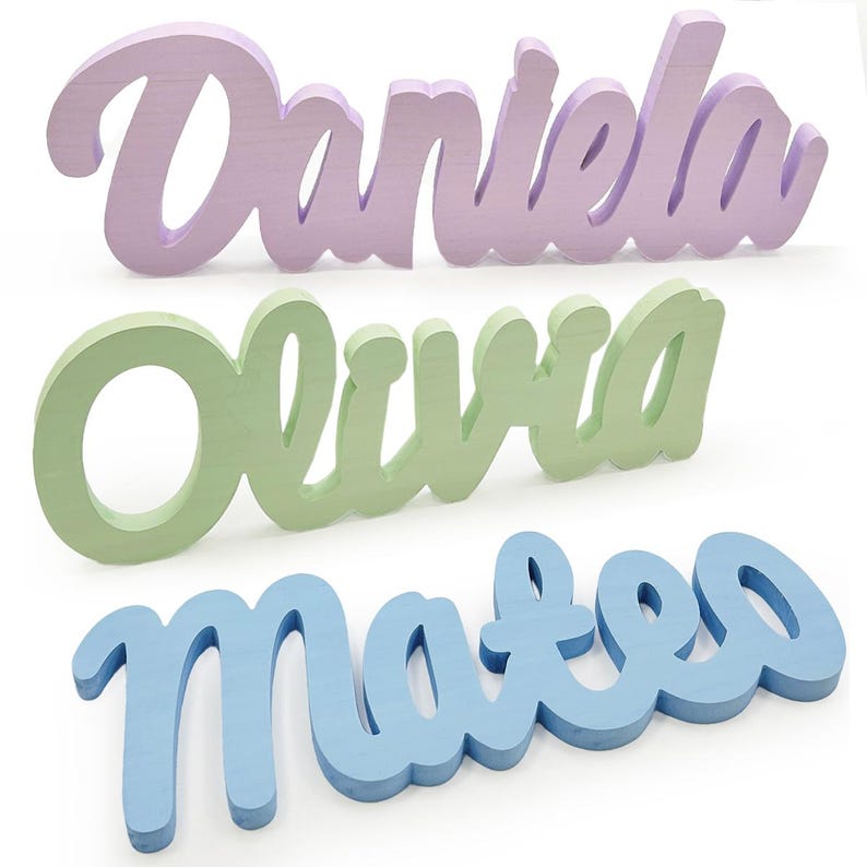 May include: Three wooden name signs in a cursive font. The names are "Daniela" in lavender, "Olivia" in light green, and "Mateo" in light blue. The signs are freestanding and painted in pastel colors.