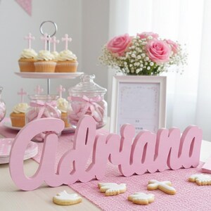 May include: A pink wooden name, "Adriana", is the focal point of a table setting. The table is decorated with cupcakes, cookies, pink roses, and a framed picture. The scene is set for a celebration.