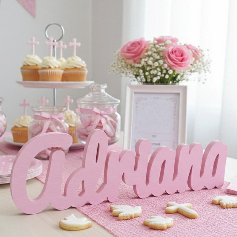 May include: A pink wooden name, "Adriana", is the focal point of a table setting. The table is decorated with cupcakes, cookies, pink roses, and a framed picture. The scene is set for a celebration.