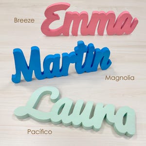 May include: Three wooden name signs in cursive script. The names are "Emma" in pink, "Martin" in blue, and "Laura" in light green. The names are displayed on a light wood-grain surface.