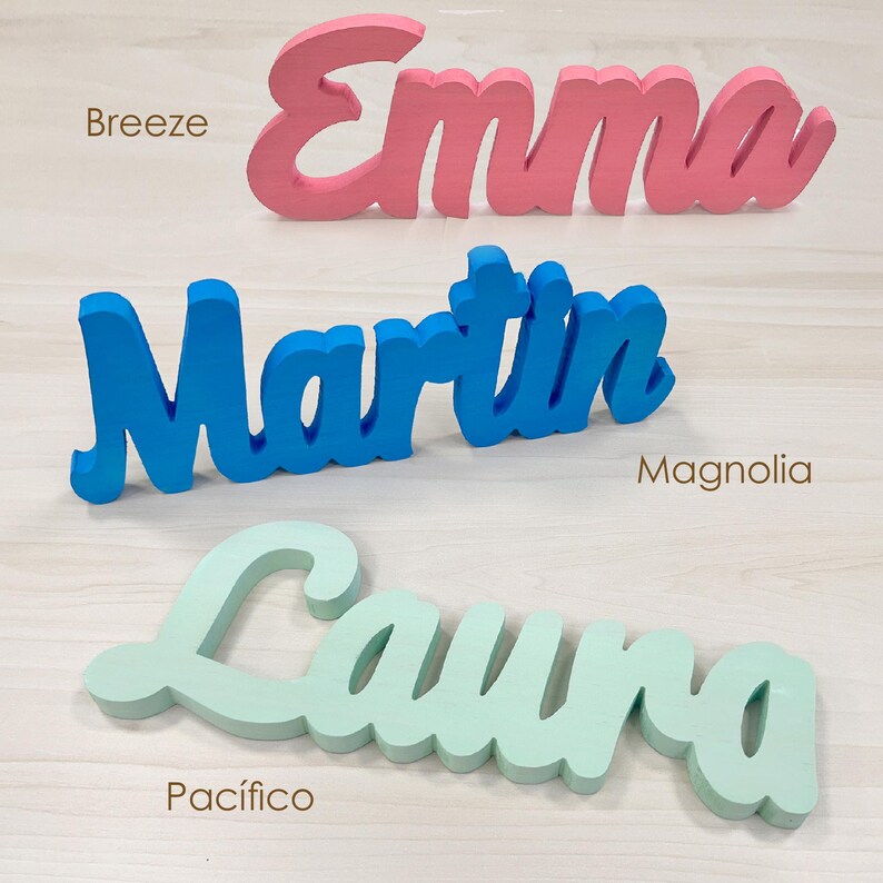 May include: Three wooden name signs in cursive script. The names are "Emma" in pink, "Martin" in blue, and "Laura" in light green. The names are displayed on a light wood-grain surface.