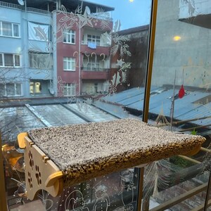 May include: A wooden cat perch with a brown fuzzy surface is mounted on a window. The window has a decorative design of a wreath with the year 2023 written inside.