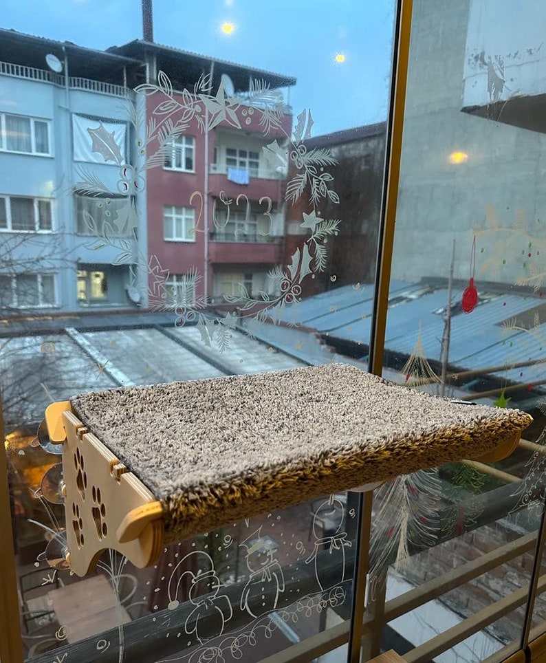 May include: A wooden cat perch with a brown fuzzy surface is mounted on a window. The window has a decorative design of a wreath with the year 2023 written inside.