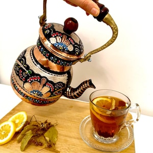 Könnte beinhalten: Eine Kupferteekanne mit einem schwarz-weißen Blumendesign und einem Holzgriff. Die Teekanne wird in eine Glas Tasse mit einer Zitronenscheibe und Tee gefüllt. Auf einem Holzbrett liegen Zitronenscheiben und getrocknete Kräuter.