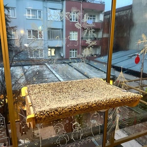 May include: A brown cat bed with a fuzzy surface is mounted on a window. The window is decorated with white drawings of snowmen and Christmas trees.