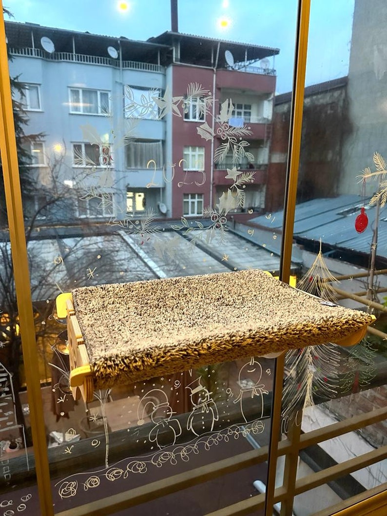 May include: A brown cat bed with a fuzzy surface is mounted on a window. The window is decorated with white drawings of snowmen and Christmas trees.