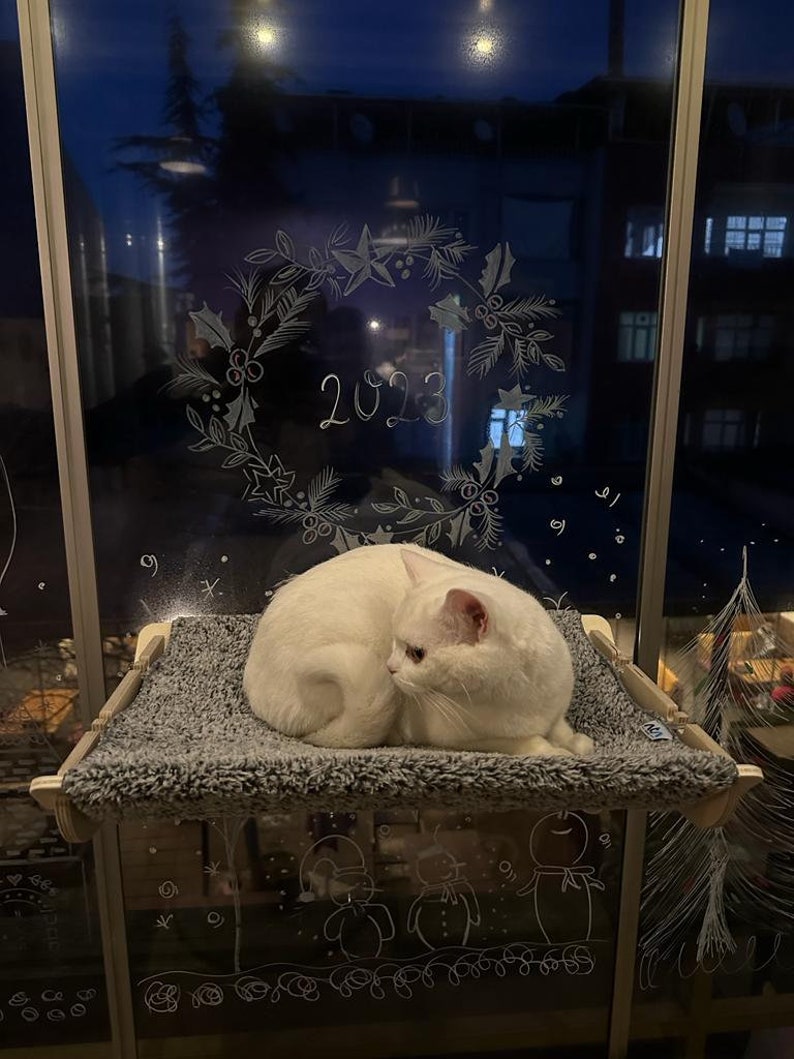 May include: A white cat is lying on a gray cat bed, which is attached to a window. The window is decorated with white chalk drawings of a wreath with the year 2023 written inside, and snowmen.