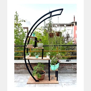 May include: A black metal plant stand with a curved design and wooden shelves. The stand has multiple shelves for displaying plants and a hanging hook for a hanging plant. The stand is outdoors on a patio.