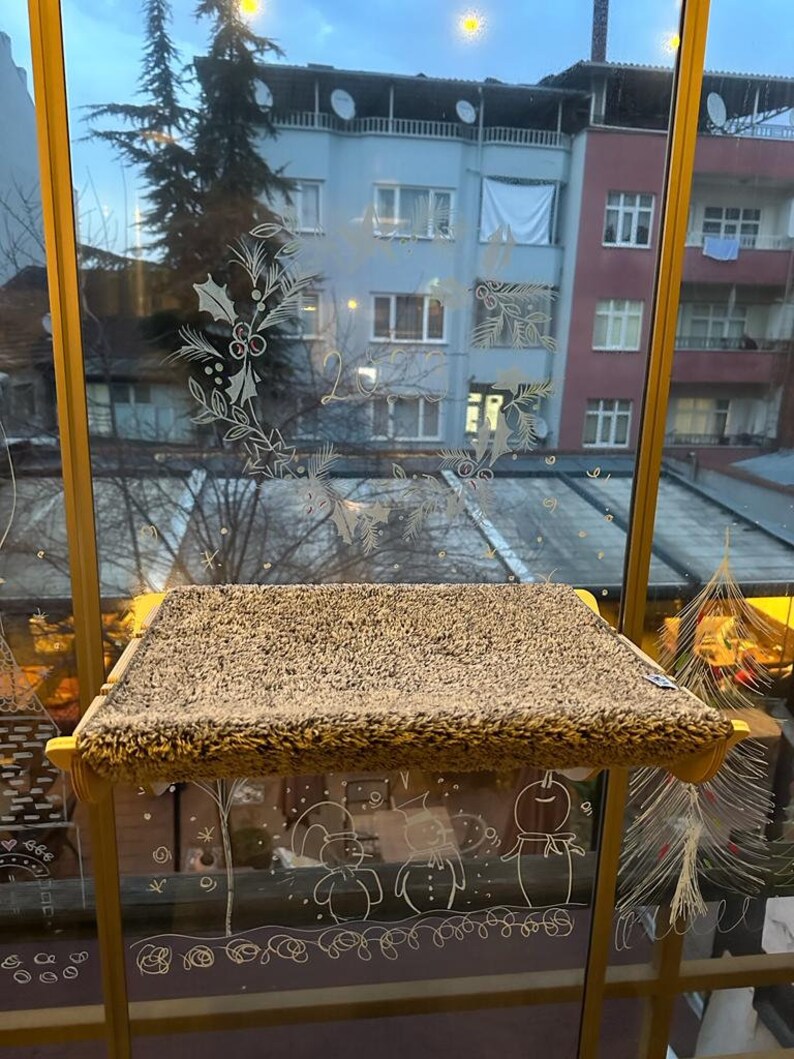 May include: A brown, fuzzy cat bed is mounted on a window with a gold frame. The window is decorated with white drawings of holly, snowmen, and a Christmas tree.