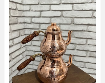 May include: A copper-colored, two-tiered teapot with floral engravings and wooden handles. The teapot has a spout and a lid. It sits on a small, brown wooden table against a white brick wall.