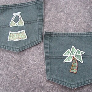 May include: Two dark olive green denim pockets featuring embroidered appliqués. One pocket displays a two-piece bikini set in light green with white stitching, while the other pocket features a palm tree design in light green with white outlines.