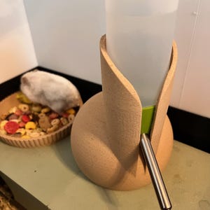 Pode incluir: Um suporte de garrafa de água de cerâmica bege para animais de estimação pequenos. O suporte tem um design curvo com uma garrafa de plástico transparente e um bico de aço inoxidável. Uma tigela de comida para animais está no fundo.