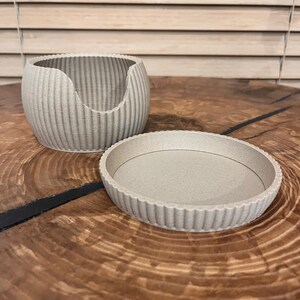 Może przedstawiać: Jasny beżowy, żebrowany ceramiczny pojemnik na rośliny z pasującym spodkiem. Doniczka ma zakrzywiony otwór z jednej strony. Spodek ma falisty brzeg. Obiekty znajdują się na drewnianej powierzchni.