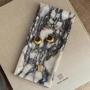 May include: A rectangular marble tray with gold hoop earrings and a gold pendant necklace. The tray has a white and gray marbled pattern. The earrings and necklace are resting on the tray.