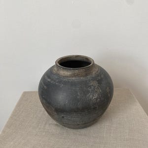 May include: A round, dark charcoal-grey ceramic vase with a wide, bulbous body and a short, thick neck. The surface features a weathered, matte texture with subtle horizontal ridges and lighter grey, worn patches around the rim and body.