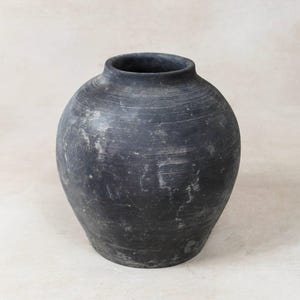 May include: A dark grey, round ceramic vase with a wide body and a narrow neck. The vase has a textured surface with visible horizontal lines and white streaks. The vase is a decorative home decor item.