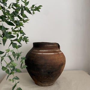 May include: A round, dark brown earthenware pot with a wide opening and a tapered base. The exterior features a weathered, textured surface with a horizontal white band encircling the upper portion of the vessel, showing signs of age and use.