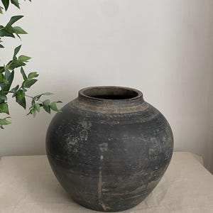 May include: A round, dark charcoal-coloured ceramic vessel with a wide, bulbous body and a short, thick neck. The surface features a matte, weathered texture with subtle horizontal striations and light grey, earthy markings across the exterior.
